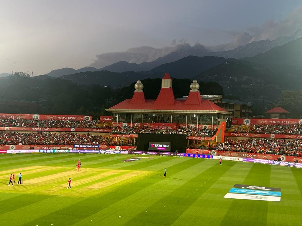 Spectators enjoying a cricket match at Dharamsala Cricket Ground, surrounded by lush green fields and panoramic Himalayan views.