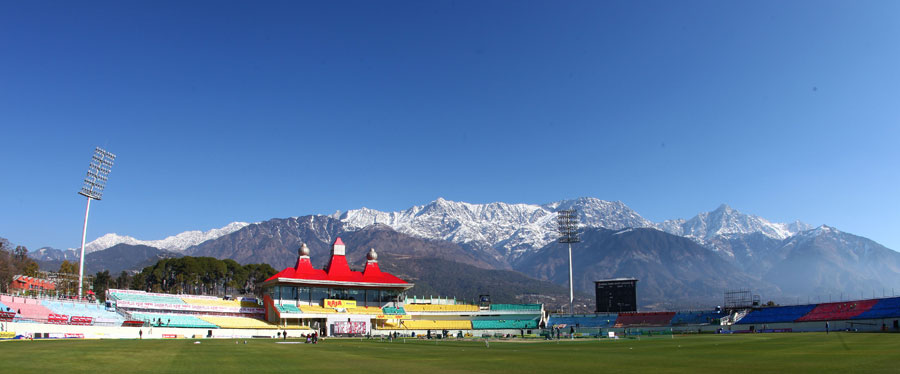 Players in action during a cricket match at Dharamsala Cricket Ground, with fans cheering in the stands and the majestic mountains looming in the distance.