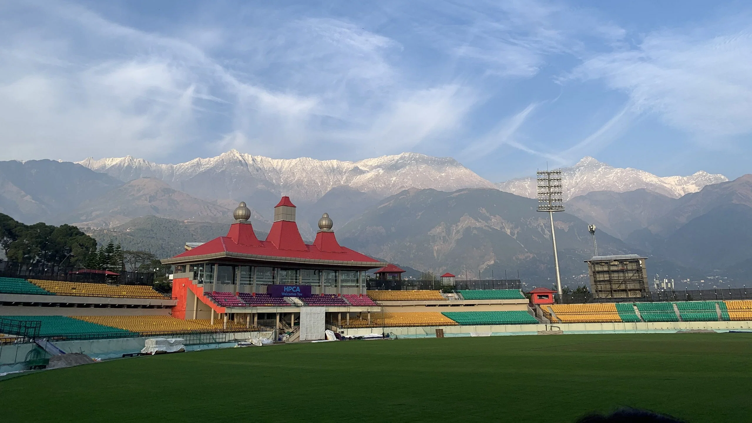 Sunset over Dharamsala Cricket Ground, casting a golden glow over the stadium and highlighting the traditional Himachali architectural style of the pavilions.
